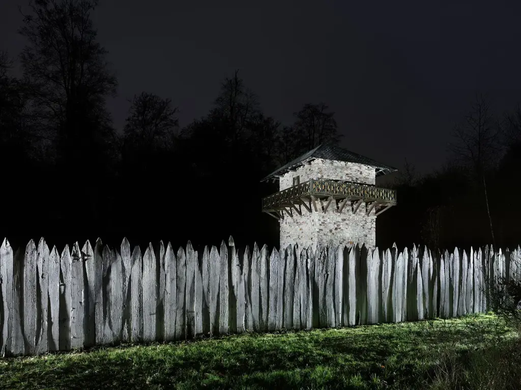 Quer durch das Bild verläuft eine Rekonstruktion des Limes mit Holzpalisade, dahinter ein Nachbau des Wachturm WP 3/15 aus Stein, beleuchtet am Abend.